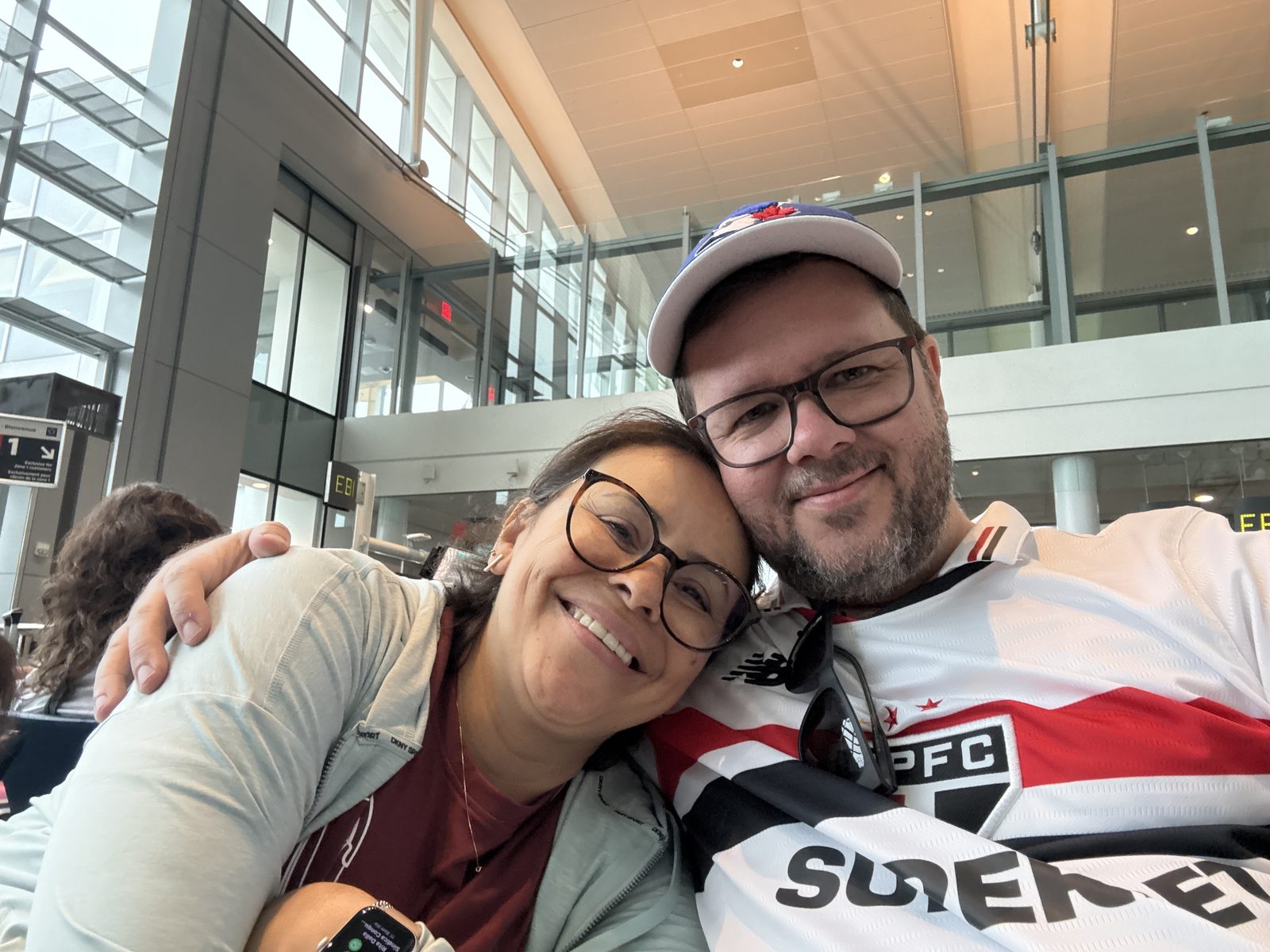 Anderson Hexsel and his partner smiling at the airport gate, Anderson wearing a São Paulo FC jersey and a Toronto Blue Jays cap